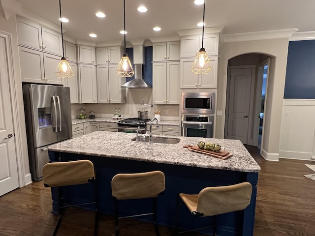 kitchen with sink, an island with sink, pendant lighting, stainless steel appliances, and wall chimney range hood