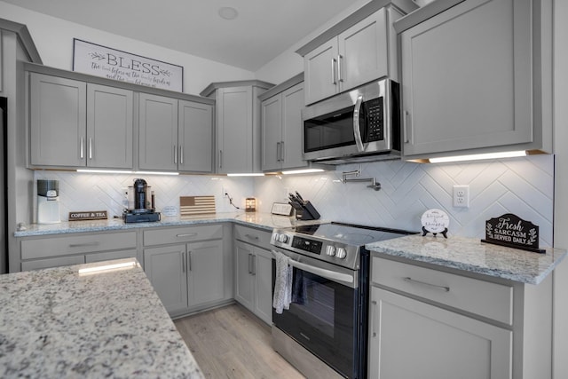 kitchen with light stone counters, light wood-type flooring, appliances with stainless steel finishes, gray cabinets, and decorative backsplash