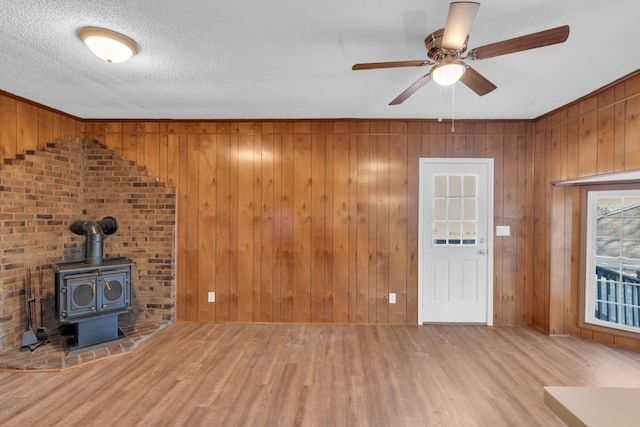 unfurnished living room with wooden walls, wood-type flooring, a wood stove, ceiling fan, and a textured ceiling