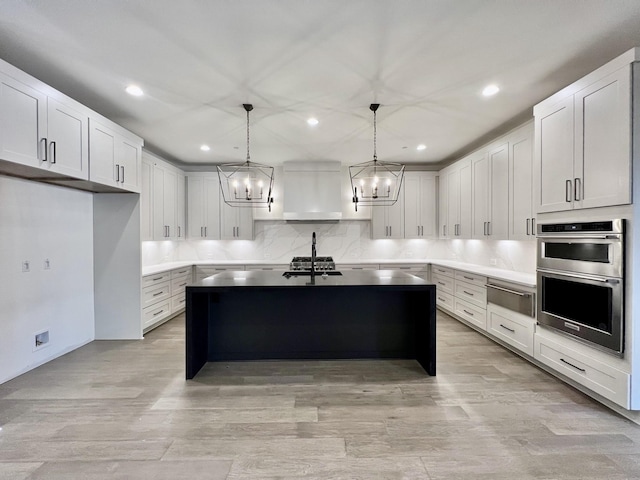 kitchen with a kitchen island with sink, stainless steel double oven, white cabinets, and decorative light fixtures