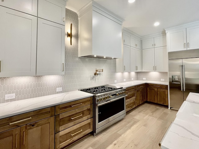 kitchen featuring wall chimney range hood, tasteful backsplash, premium appliances, light stone countertops, and white cabinets