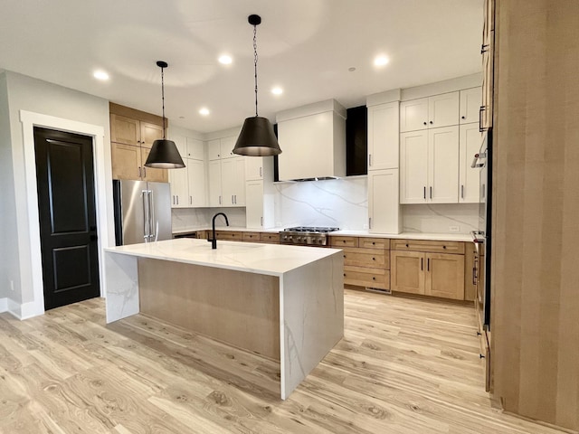 kitchen with appliances with stainless steel finishes, pendant lighting, a kitchen island with sink, wall chimney range hood, and light hardwood / wood-style flooring