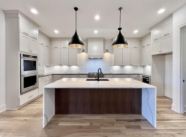 kitchen featuring hanging light fixtures, an island with sink, stainless steel double oven, light stone countertops, and wall chimney range hood