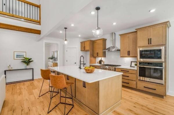 kitchen with an island with sink, wall chimney range hood, light wood-type flooring, and decorative light fixtures