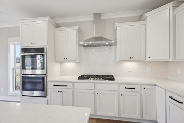 kitchen with appliances with stainless steel finishes, white cabinetry, backsplash, ornamental molding, and wall chimney range hood