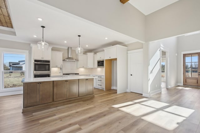 kitchen with wall chimney range hood, white cabinetry, stainless steel appliances, an island with sink, and decorative light fixtures