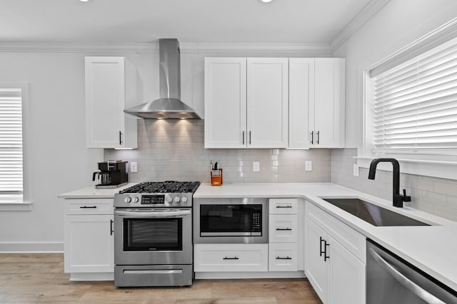 kitchen with appliances with stainless steel finishes, sink, white cabinets, ornamental molding, and wall chimney range hood