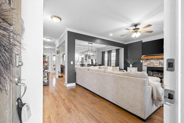 living room with crown molding, a stone fireplace, ceiling fan with notable chandelier, and light wood-type flooring