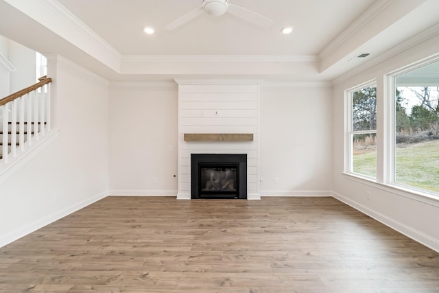 unfurnished living room with a large fireplace, ceiling fan, a tray ceiling, crown molding, and light wood-type flooring