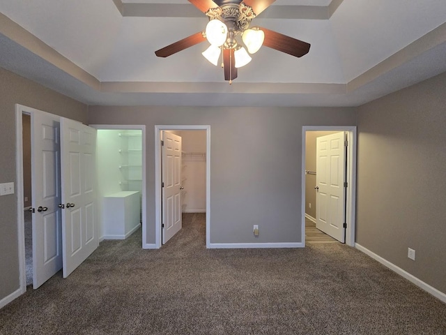 unfurnished bedroom featuring ceiling fan, a spacious closet, a raised ceiling, and dark colored carpet