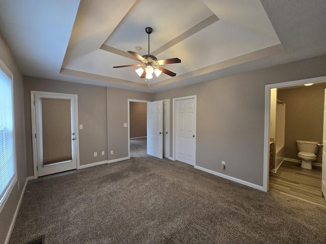 unfurnished bedroom with dark colored carpet, ensuite bathroom, ceiling fan, and a tray ceiling