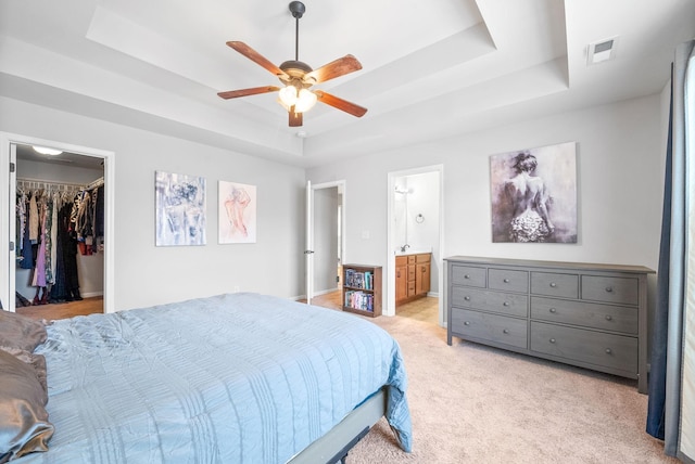 bedroom featuring connected bathroom, a spacious closet, light colored carpet, a raised ceiling, and ceiling fan