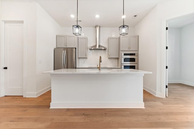 kitchen with wall chimney exhaust hood, sink, hanging light fixtures, an island with sink, and stainless steel appliances