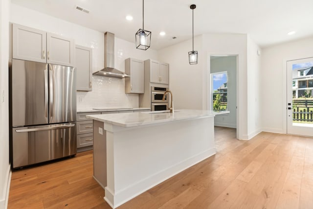 kitchen featuring a kitchen island with sink, wall chimney range hood, gray cabinets, and appliances with stainless steel finishes