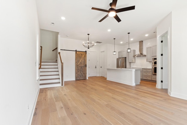 unfurnished living room featuring a barn door, ceiling fan with notable chandelier, and light hardwood / wood-style floors