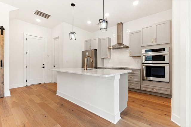 kitchen featuring appliances with stainless steel finishes, gray cabinetry, a kitchen island with sink, a barn door, and wall chimney range hood