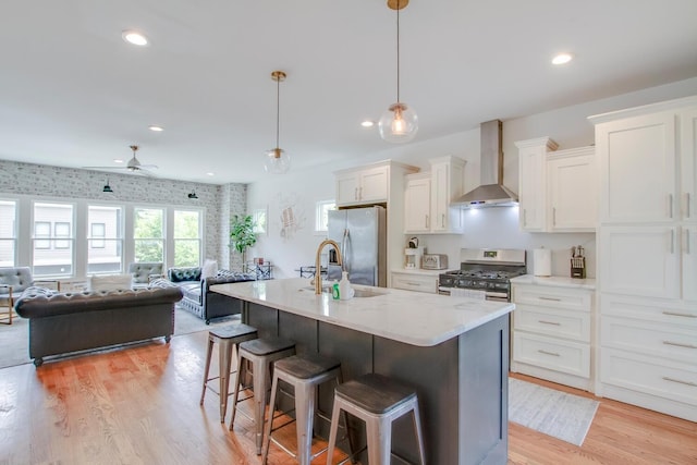 kitchen with pendant lighting, appliances with stainless steel finishes, an island with sink, white cabinets, and wall chimney exhaust hood