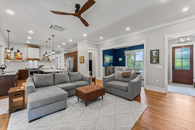 living room with crown molding, ceiling fan with notable chandelier, sink, and light wood-type flooring