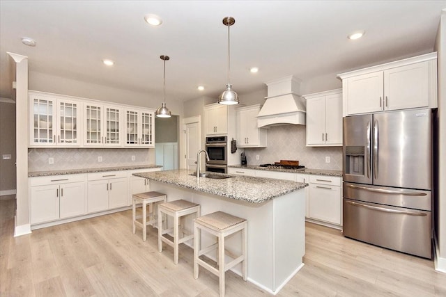 kitchen with appliances with stainless steel finishes, white cabinets, custom exhaust hood, light stone counters, and a center island with sink