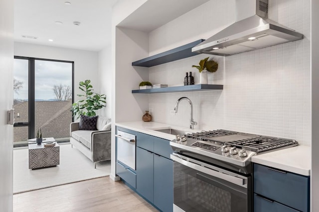 kitchen featuring wall chimney range hood, sink, light hardwood / wood-style flooring, stainless steel gas range, and blue cabinets