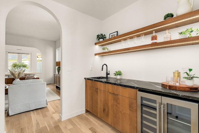 bar featuring beverage cooler, sink, light hardwood / wood-style flooring, and dark stone countertops