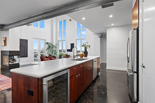 kitchen with sink, a center island with sink, a towering ceiling, stainless steel appliances, and beverage cooler