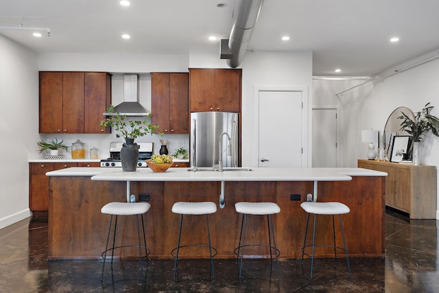 kitchen with stainless steel fridge, gas range oven, an island with sink, and wall chimney range hood