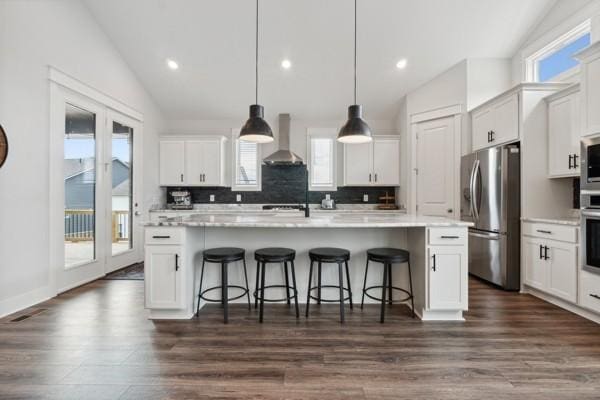 kitchen with pendant lighting, appliances with stainless steel finishes, white cabinetry, a center island with sink, and wall chimney exhaust hood