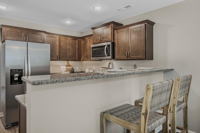 kitchen with a breakfast bar, dark brown cabinets, kitchen peninsula, stainless steel appliances, and light stone countertops