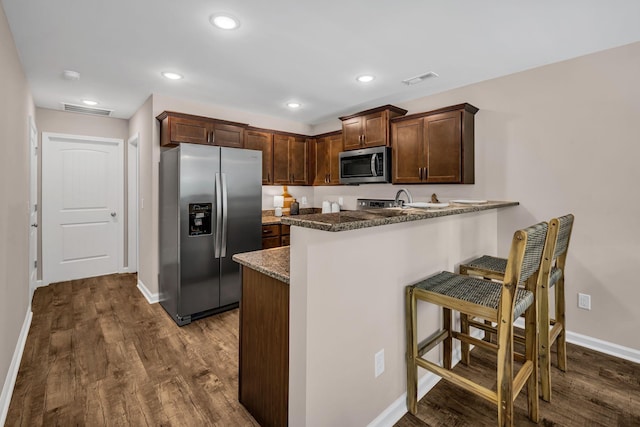 kitchen featuring appliances with stainless steel finishes, a kitchen bar, dark hardwood / wood-style flooring, dark stone counters, and kitchen peninsula