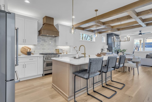 kitchen featuring a center island with sink, hanging light fixtures, custom range hood, stainless steel appliances, and white cabinets