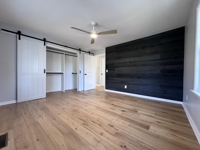 unfurnished bedroom with ceiling fan, a barn door, wooden walls, and light hardwood / wood-style floors