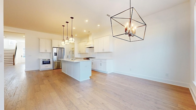 kitchen featuring stainless steel refrigerator with ice dispenser, white cabinetry, an island with sink, custom range hood, and pendant lighting