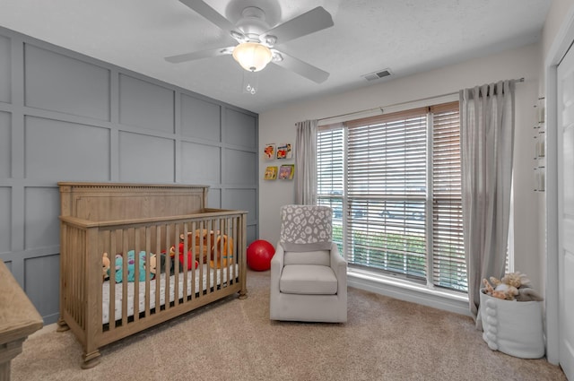 bedroom featuring light carpet, visible vents, a ceiling fan, a nursery area, and a decorative wall