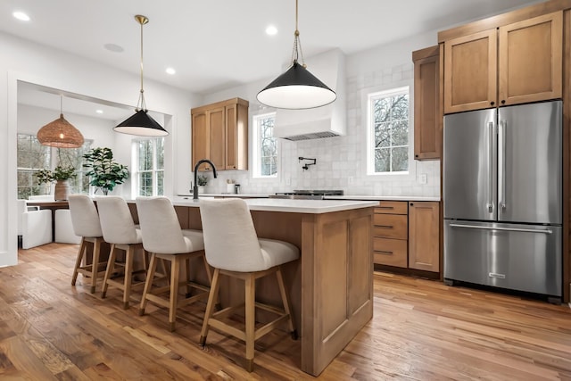 kitchen featuring high quality fridge, decorative light fixtures, an island with sink, and light wood-type flooring