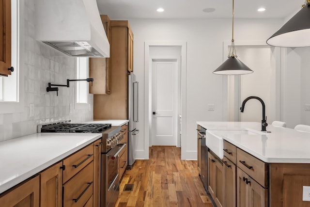 kitchen with hanging light fixtures, light hardwood / wood-style flooring, appliances with stainless steel finishes, an island with sink, and range hood