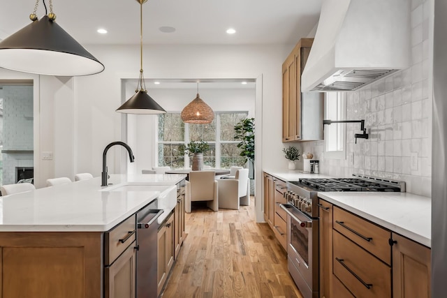 kitchen featuring appliances with stainless steel finishes, light hardwood / wood-style floors, custom range hood, an island with sink, and decorative light fixtures