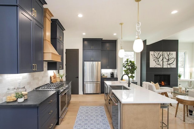 kitchen with a kitchen island with sink, stainless steel appliances, a sink, dark countertops, and decorative light fixtures