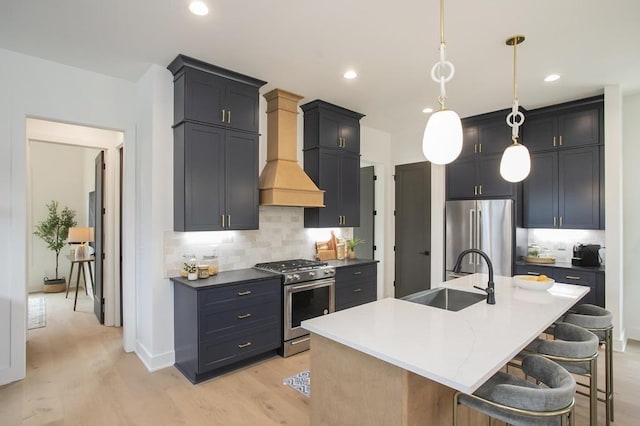 kitchen featuring a kitchen island with sink, a sink, appliances with stainless steel finishes, decorative light fixtures, and custom range hood