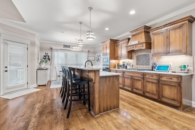 kitchen featuring premium range hood, decorative light fixtures, an island with sink, light hardwood / wood-style floors, and stainless steel appliances