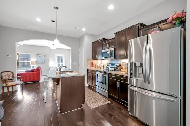 kitchen featuring dark wood-type flooring, a breakfast bar area, appliances with stainless steel finishes, light stone countertops, and an island with sink