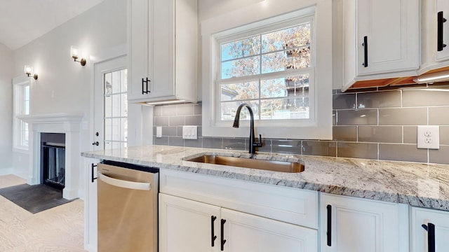 kitchen featuring light stone counters, stainless steel dishwasher, a glass covered fireplace, white cabinets, and a sink