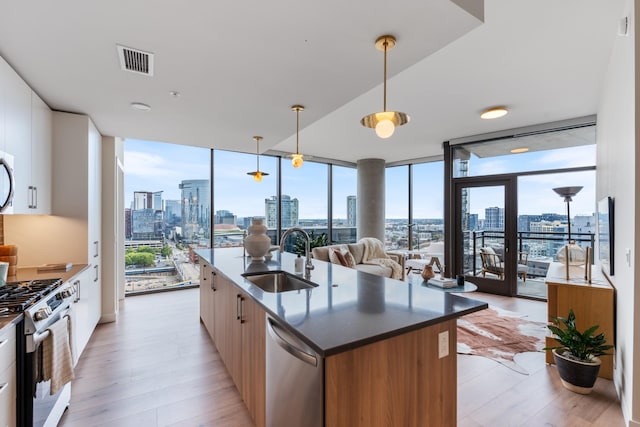 kitchen with sink, hanging light fixtures, appliances with stainless steel finishes, an island with sink, and a wall of windows