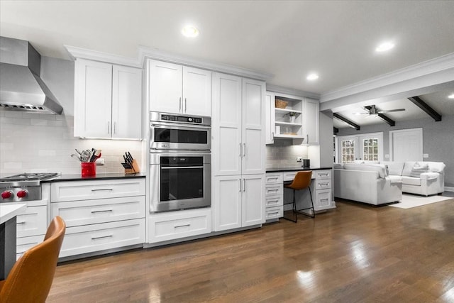 kitchen with white cabinetry, stainless steel appliances, dark hardwood / wood-style floors, built in desk, and wall chimney exhaust hood