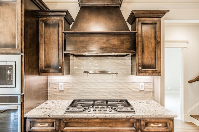 kitchen with light stone counters, custom exhaust hood, decorative backsplash, appliances with stainless steel finishes, and dark brown cabinets