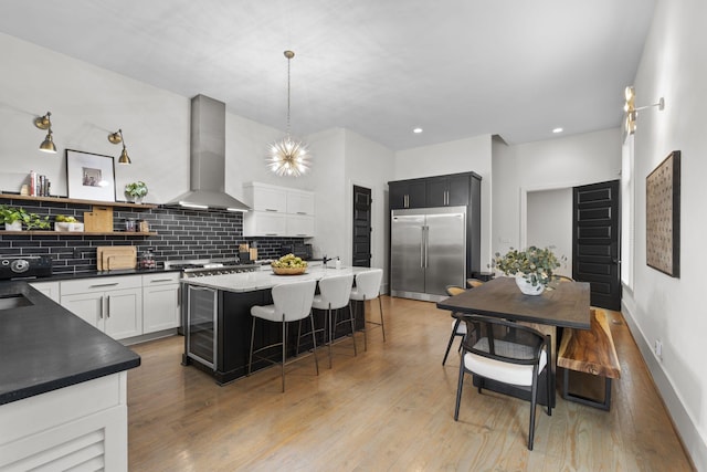 kitchen with pendant lighting, wall chimney range hood, a breakfast bar, white cabinetry, and built in fridge