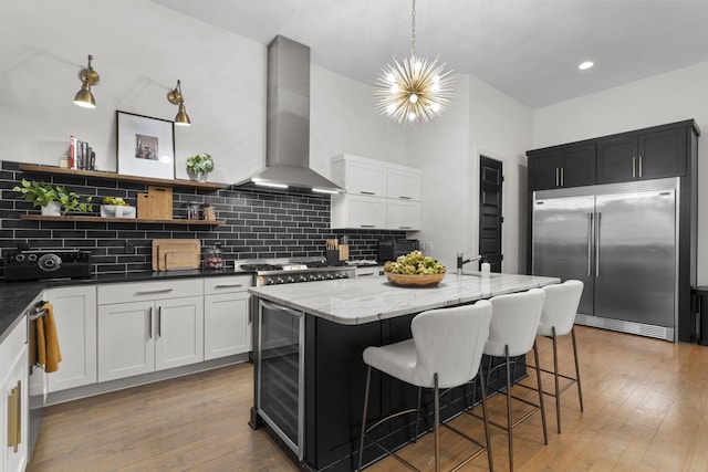 kitchen with appliances with stainless steel finishes, decorative light fixtures, dark stone countertops, a center island with sink, and wall chimney exhaust hood