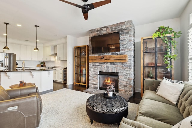 living room with wine cooler, a stone fireplace, sink, dark hardwood / wood-style flooring, and ceiling fan