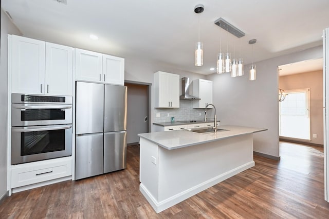 kitchen featuring wall chimney exhaust hood, white cabinetry, an island with sink, pendant lighting, and stainless steel appliances