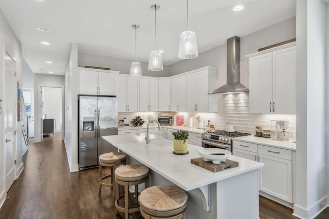 kitchen with appliances with stainless steel finishes, decorative light fixtures, white cabinets, a kitchen island with sink, and wall chimney range hood